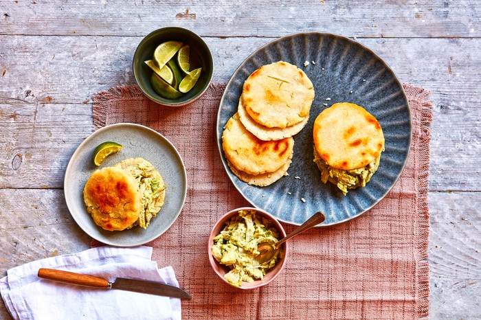 a plate of arepas with a small bowl of lime wedges and a knife on a pink fabric background