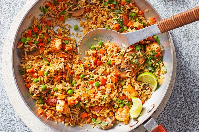 A silver pan of rice with vegetables with a serving spoon on a stone board next to wedges of lime