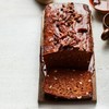 A dark brown Date and pecan loaf cake with chocolate bitters on a small board with a white background