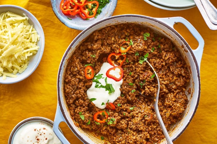 A casserole dish of chilli topped with sour cream next to bowls of grated cheese and coriander