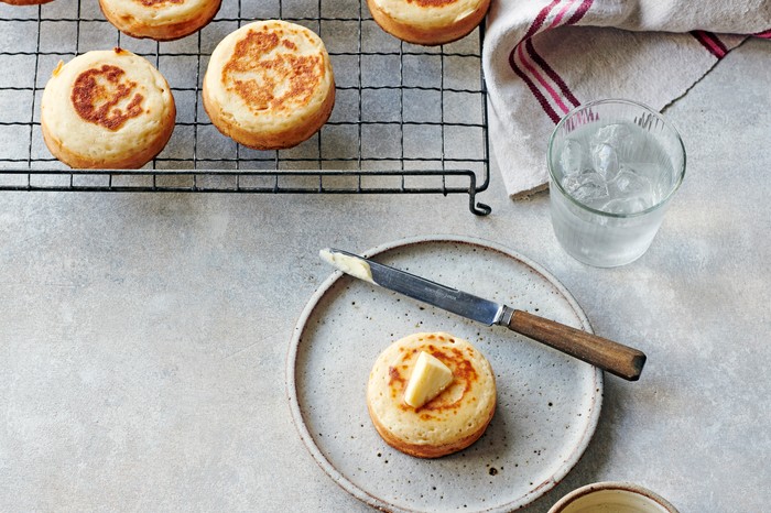 A wire cooling rack filled with golden crumpets, with one crumpet on a plate with a knife and butter