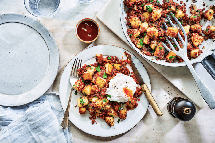 Two white plates of golden fried potatoes topped with fried egg on a white wooden surface with a knife and fork and pot of brown sauce